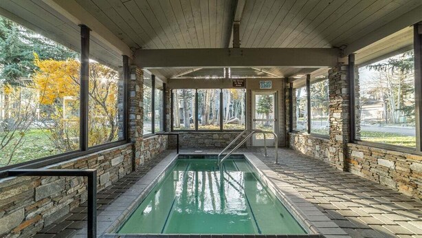 Una piscina interior con paredes de piedra y techo de madera, rodeada por amplios ventanales.