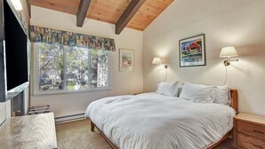A bedroom with a large bed, two bedside lamps, a window with a view of trees, and a television on the left. - Chateau Blanc 30: One car garage, near town (Mammoth Lakes)