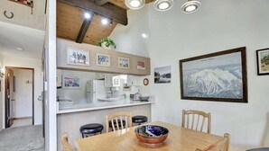 A dining area with a wooden table, chairs, and a large bowl. There is a kitchen area with a counter and various appliances. The room has wooden beams on the ceiling and a framed picture on the wall. - Chateau Blanc 30: One car garage, near town (Mammoth Lakes)