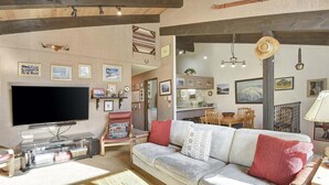 A living room with a large flat-screen TV, a grey sofa with red and white cushions, wooden beams on the ceiling, and framed pictures on the walls. - Chateau Blanc 30: One car garage, near town (Mammoth Lakes)