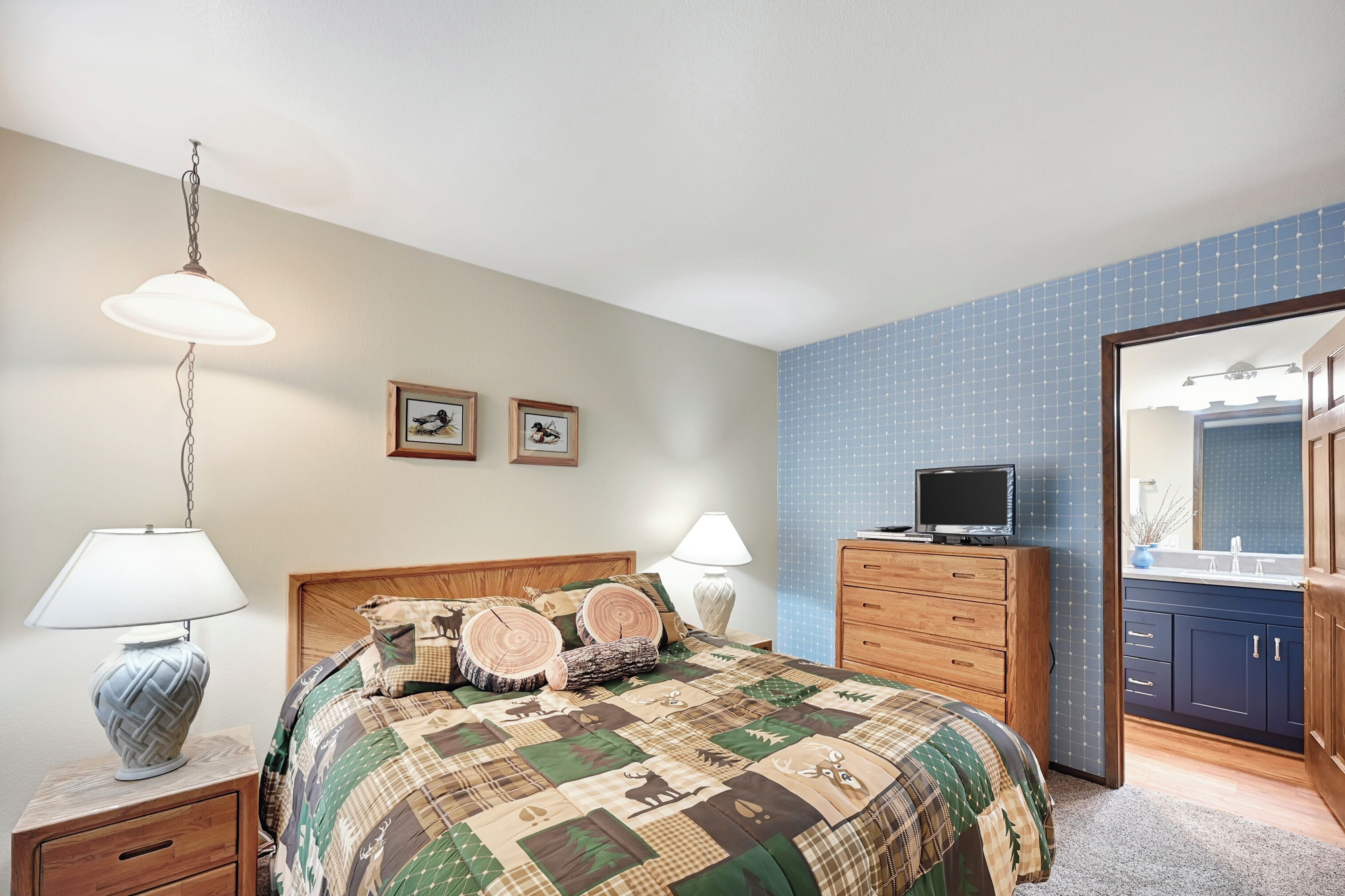 A bedroom with a bed, bedside lamps, a dresser, a television, and a bathroom with a mirror and sink.