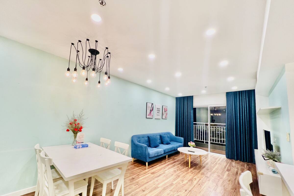 Ein modernes Wohnzimmer mit einem blauen Sofa, einem weißen Esstisch, Stühlen, einer Chandelüster und einem Balkon mit blauen Vorhängen.