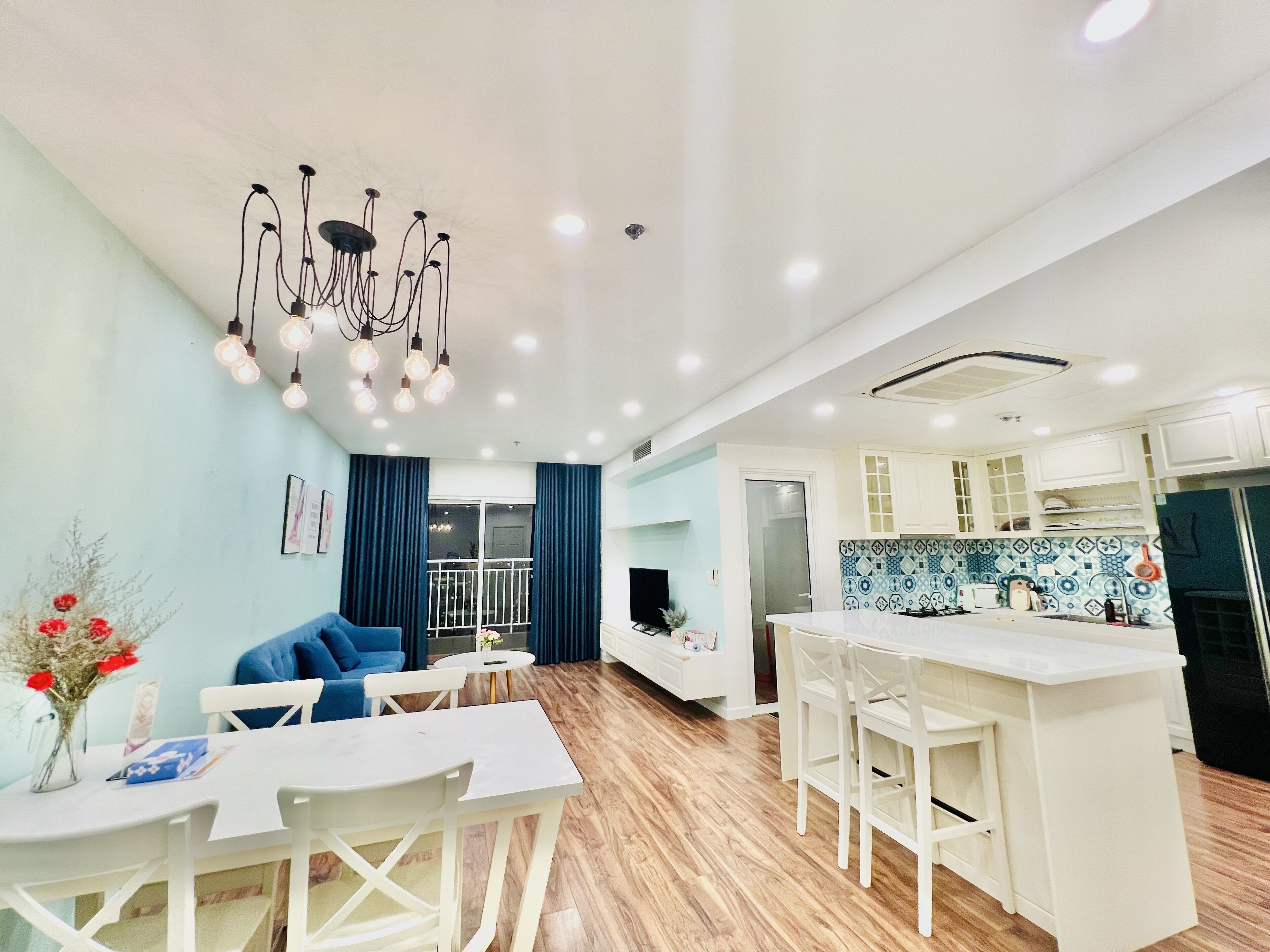 A modern living room with a blue sofa, white dining table, and a kitchen area with white cabinets and a blue backsplash.