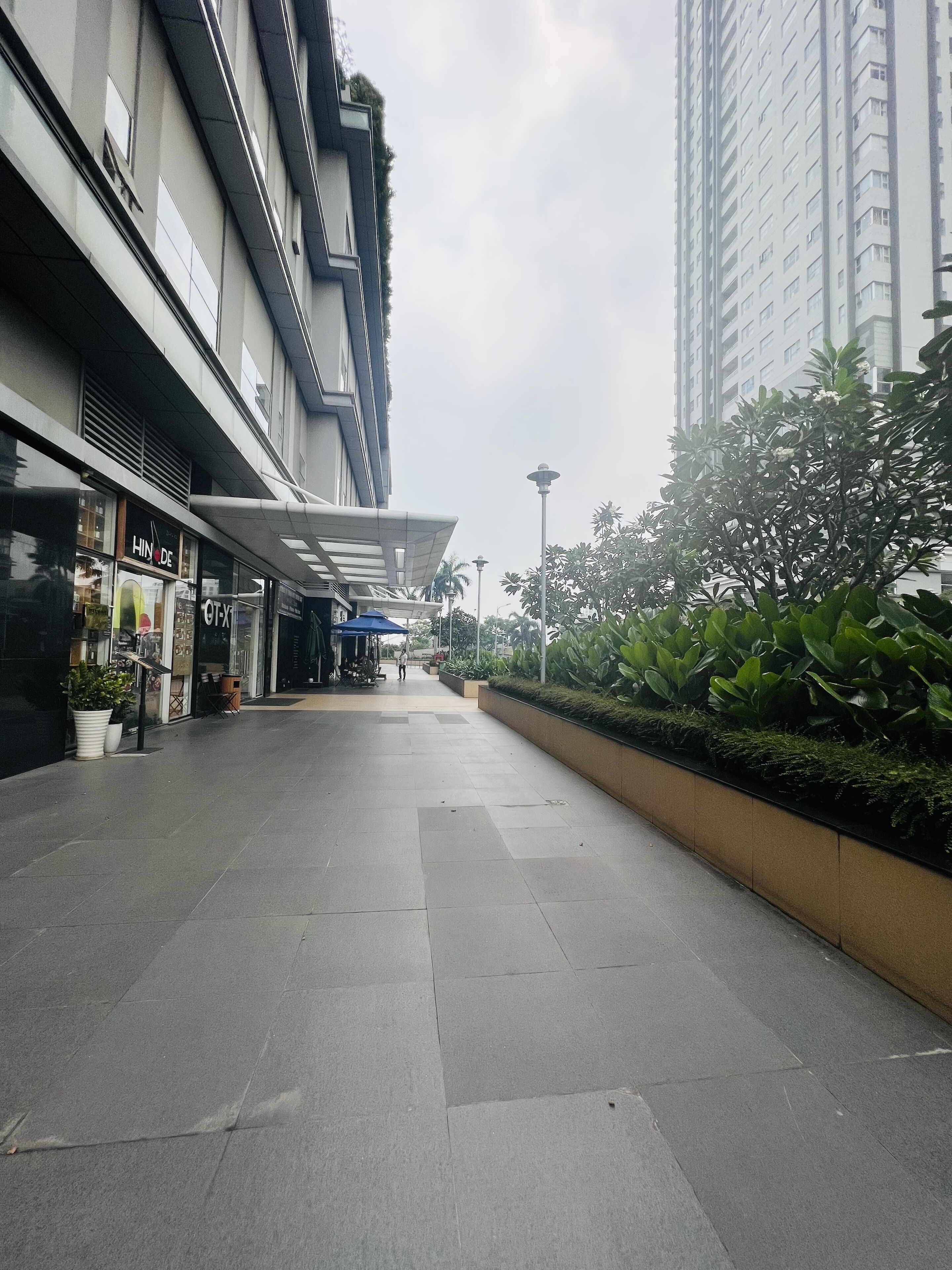 A modern urban walkway with a storefront on the left, a planter with greenery on the right, and a tall building in the background.