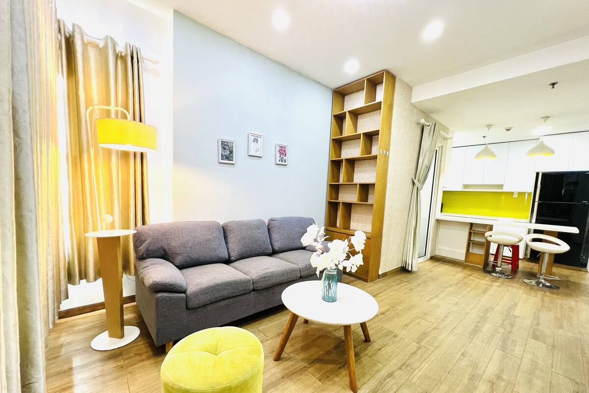 Ein modernes Wohnzimmer mit einem grauen Sofa, einem gelben Pouf, einem weißen runden Tisch mit Blumen und einer Holzhockerreihung.