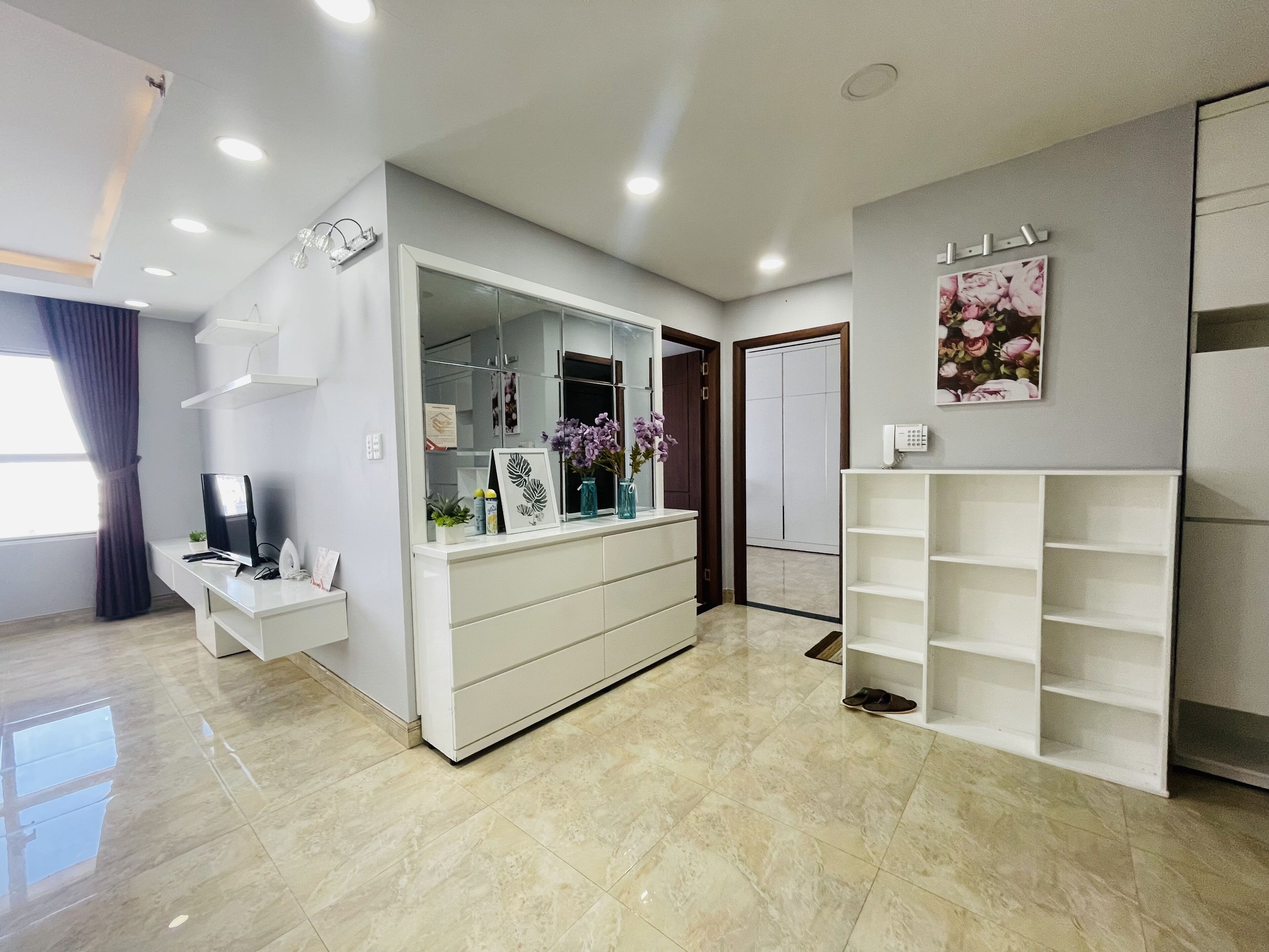 Een moderne, goed verlichte kamer met een wit bureau, een computer en een spiegel. Er is een garderobe met spiegel, een plank met decoratieve items, en een deur naar een andere kamer.