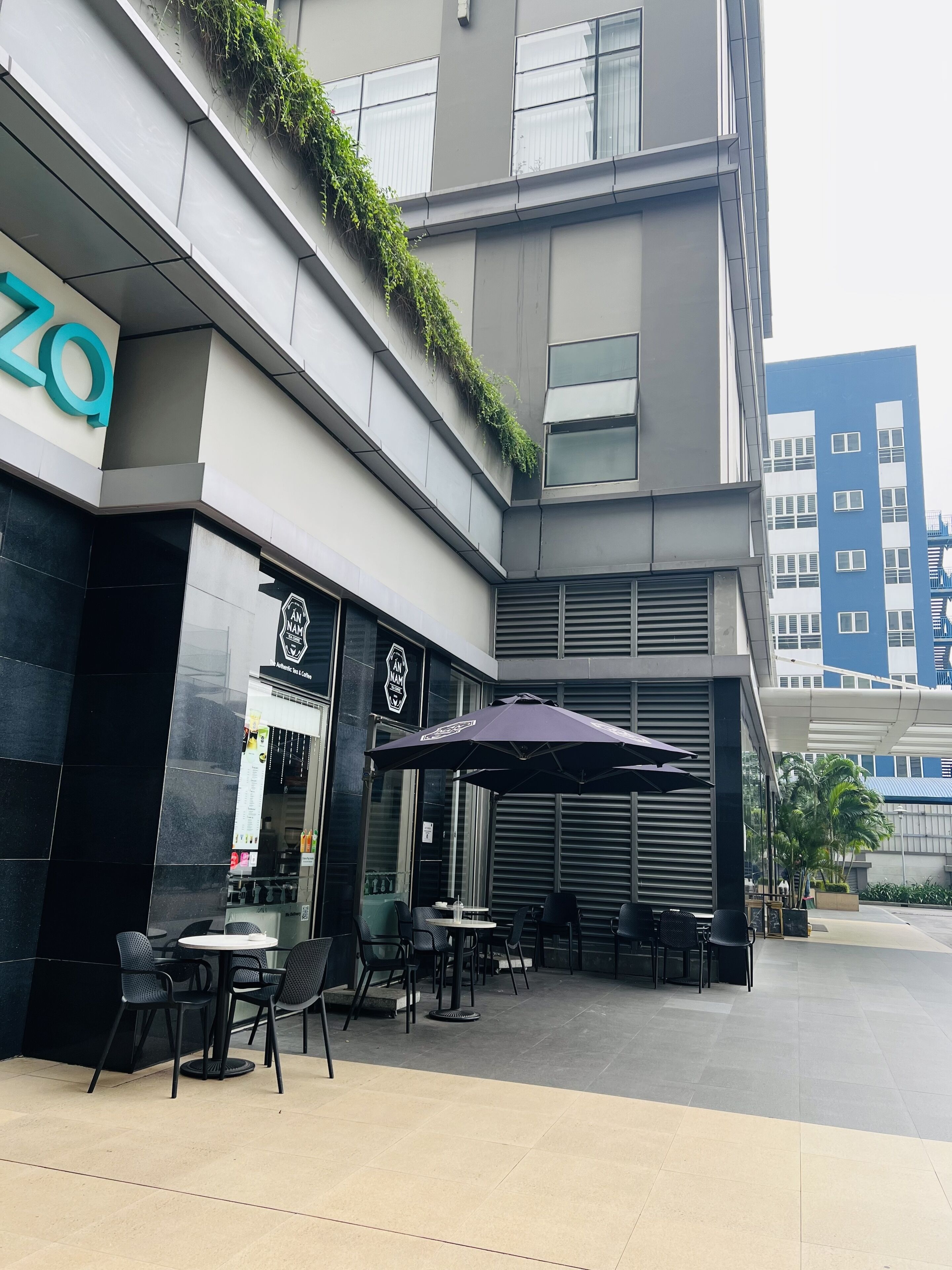 An outdoor cafe area with tables and chairs, a large umbrella, and a building with a '20' sign and a logo.
