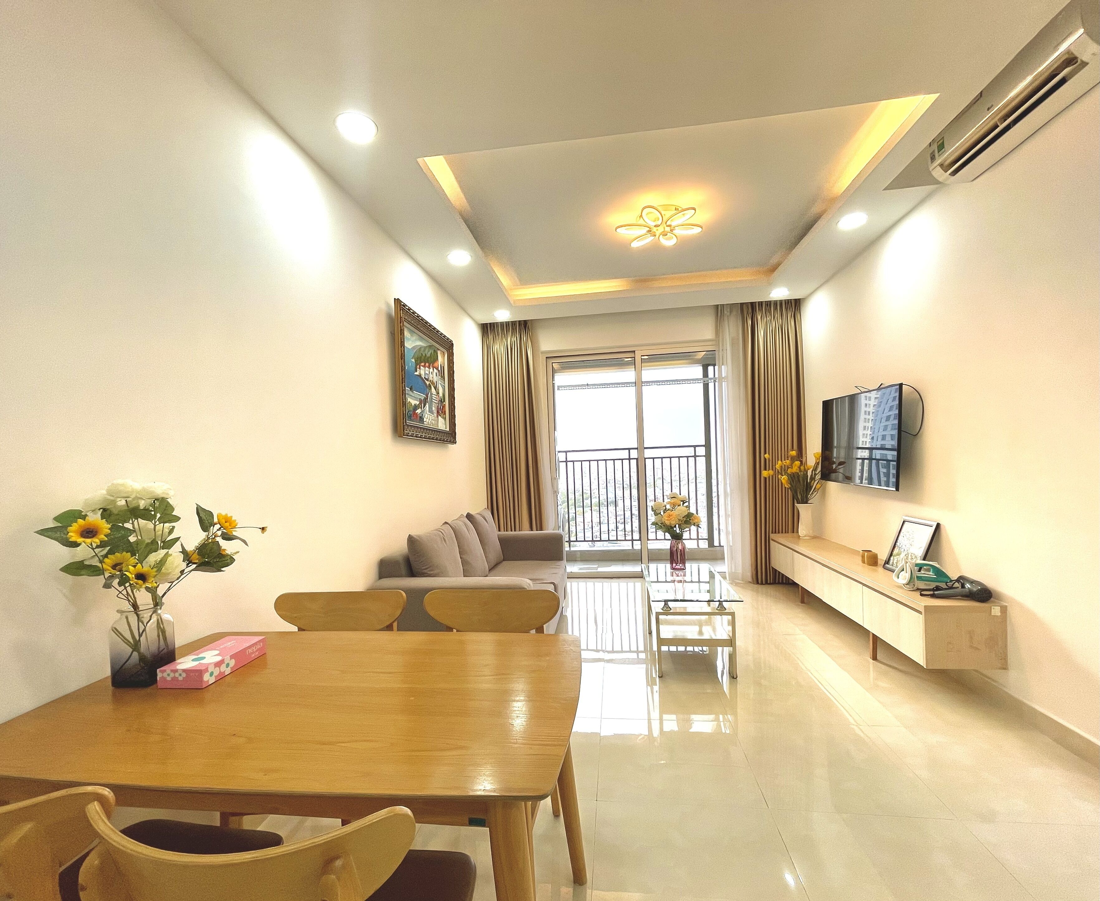 A modern living room with a dining area, a sofa, a TV mounted on the wall, and a balcony with curtains.