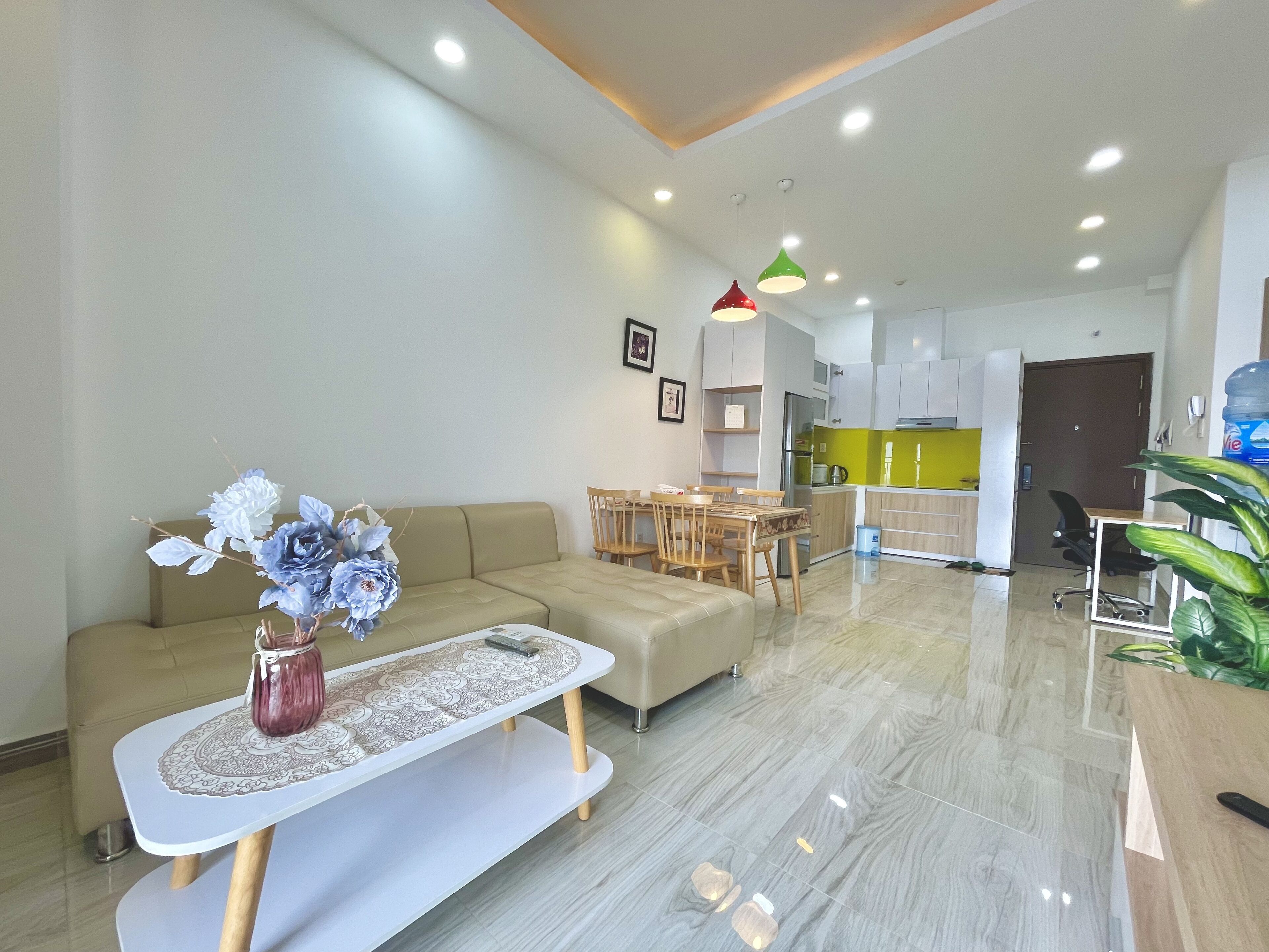 A modern living room with a sofa, a coffee table, and a vase of flowers. There is a dining area with a table and chairs, and a kitchen in the background.