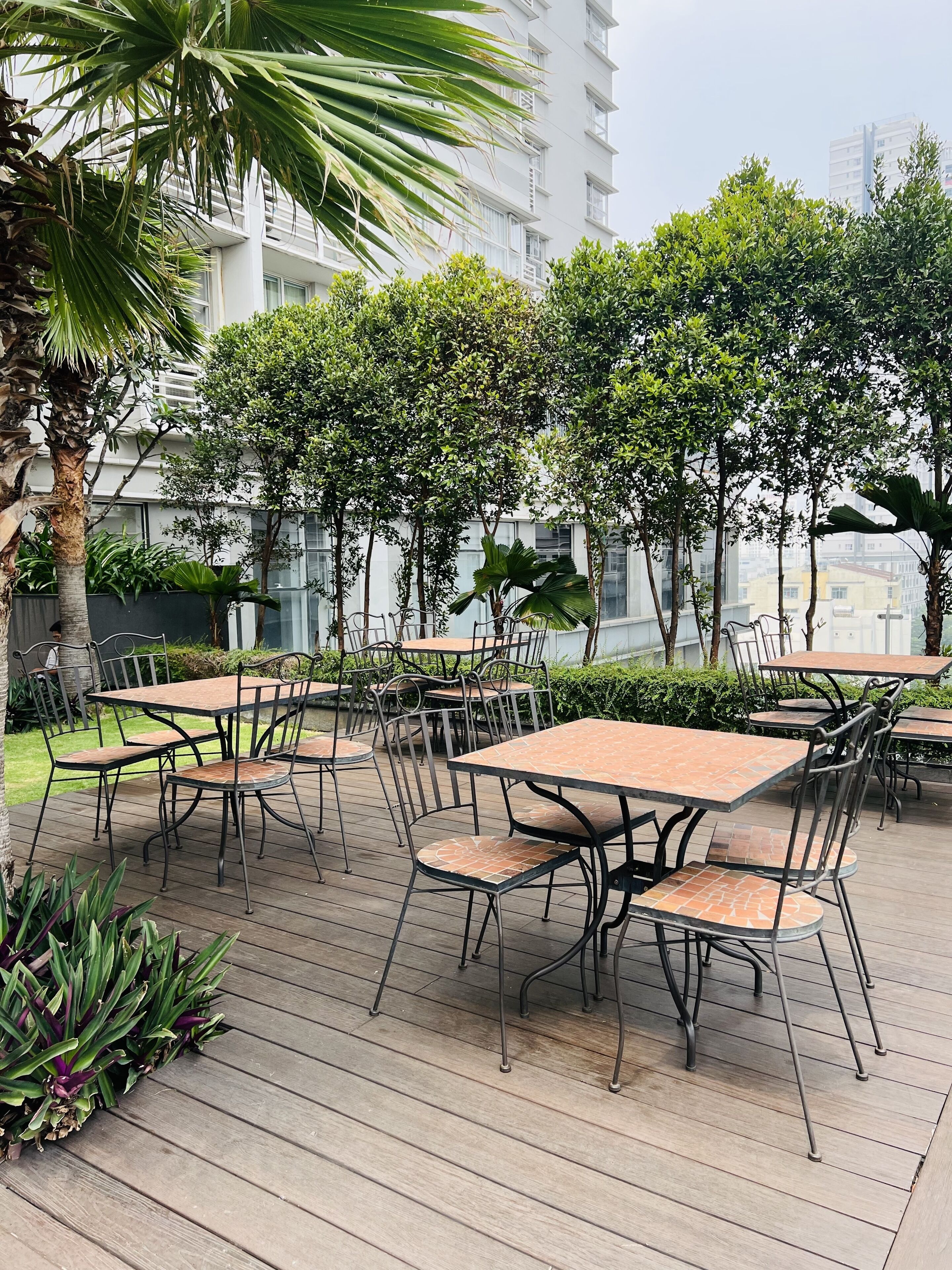 An outdoor patio area with multiple tables and chairs, surrounded by greenery and buildings in the background.