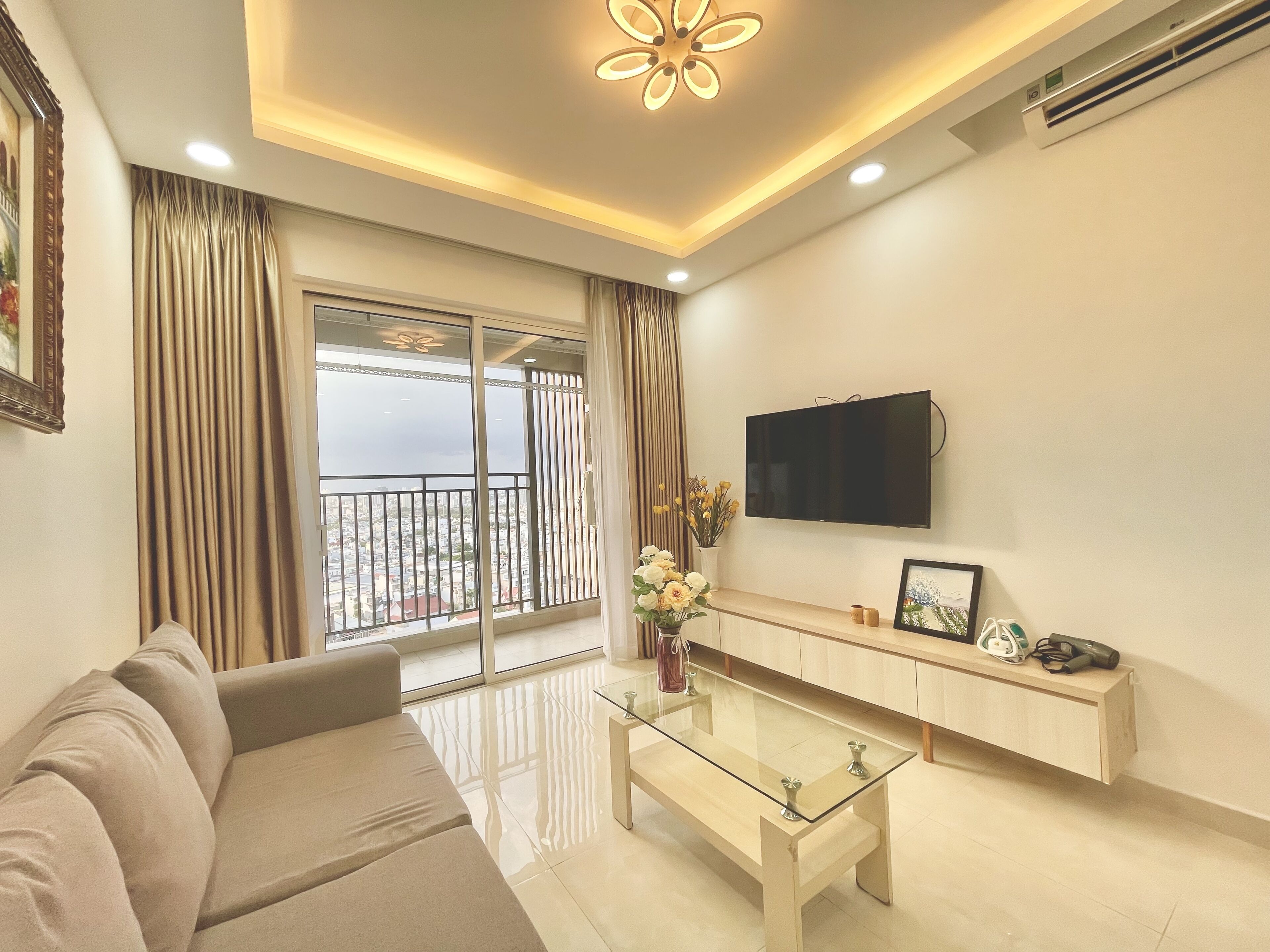 A modern living room with a sofa, a glass coffee table, a flat-screen TV mounted on the wall, and a balcony with a view.