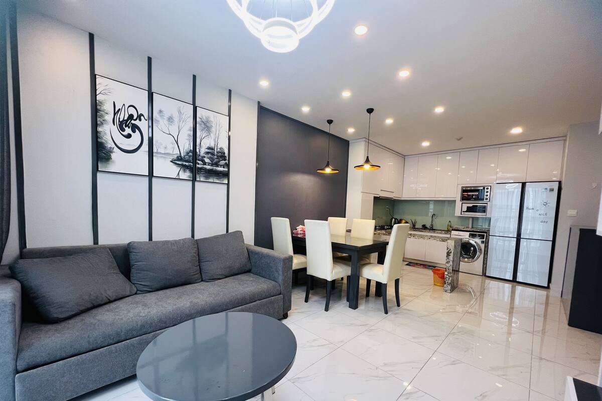 Ein modernes Wohnzimmer mit grauem Sofa, einem runden Couchtisch und einem Essbereich mit weißem Tisch und Stühlen. Im Hintergrund ist eine Küche mit weißen Schränken und einem Einbauofen zu sehen.