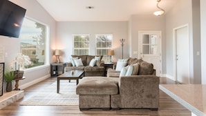 A spacious living room with a large sectional sofa, a coffee table, and two lamps. There is a flat-screen TV mounted on the wall, and a large window providing natural light. - Fabulous 4 Bed/3 Bath Rental on Lodge Hill! Updated, Charming & Pet-Friendly! (Cambria)