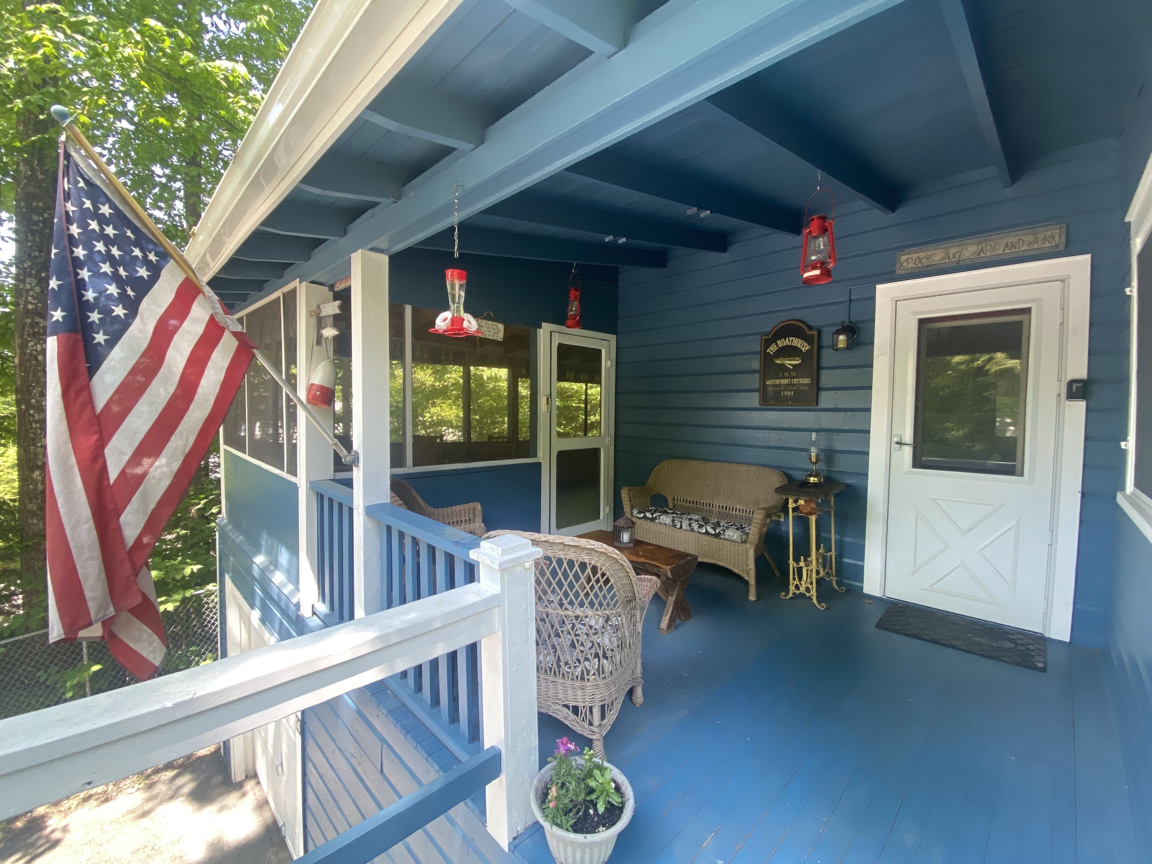 Eine Veranda mit blauer Geländer, ein Korbstuhl, ein Blumentopf und eine amerikanische Flagge.