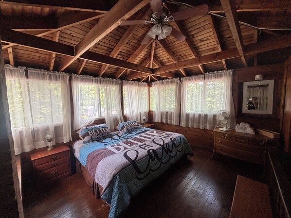 A wooden cabin bedroom with a bed, two bedside tables, a ceiling fan, and a window with curtains.