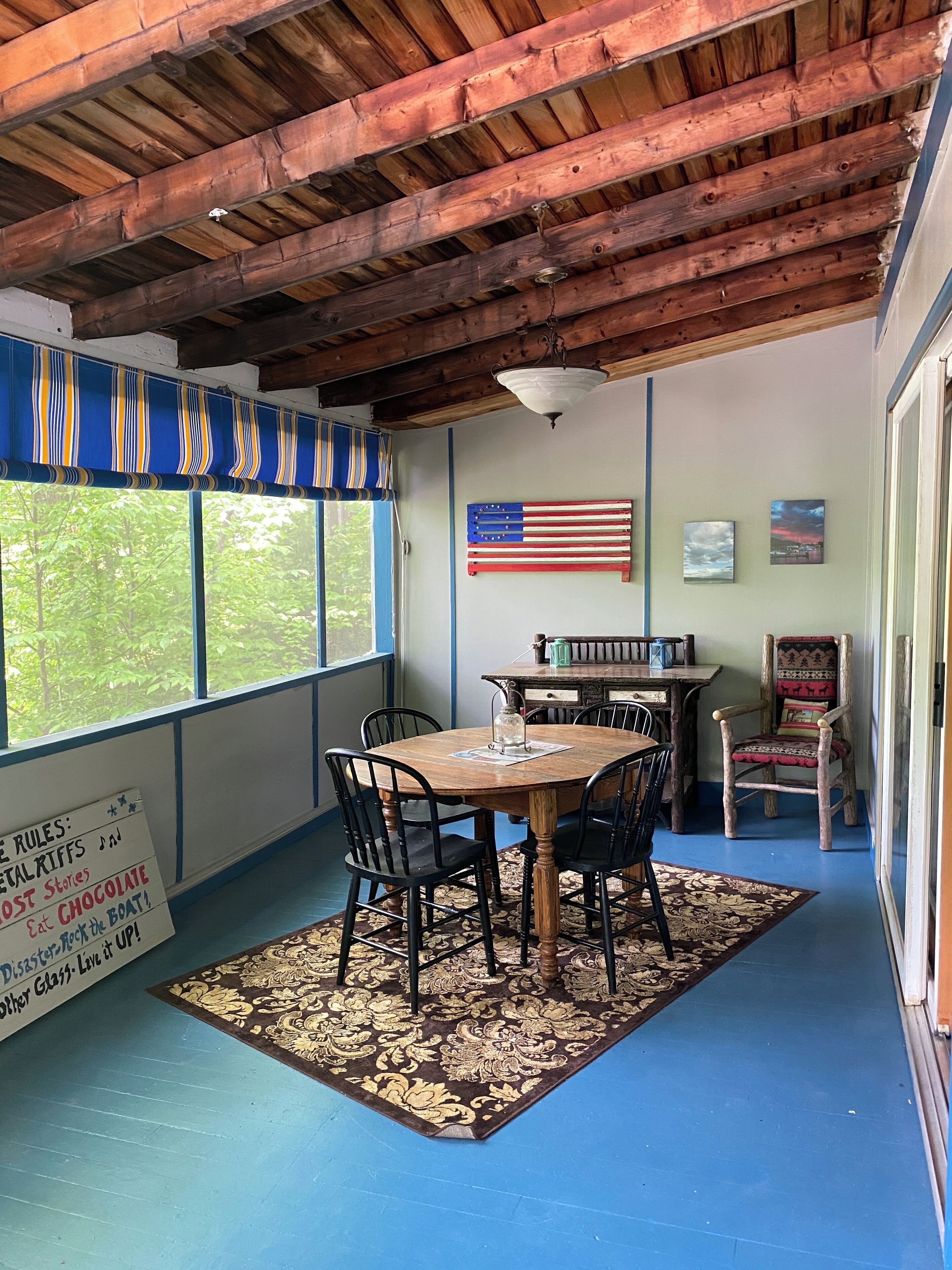 A covered patio area with a wooden table and chairs, a flag on the wall, and a view of greenery outside.