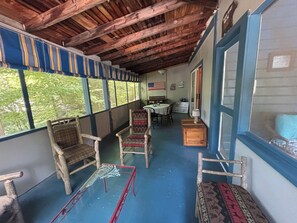 A covered porch with wooden chairs, a table, and a view of greenery through large windows. - Charming Adirondack Style Summer Home (Lake George)