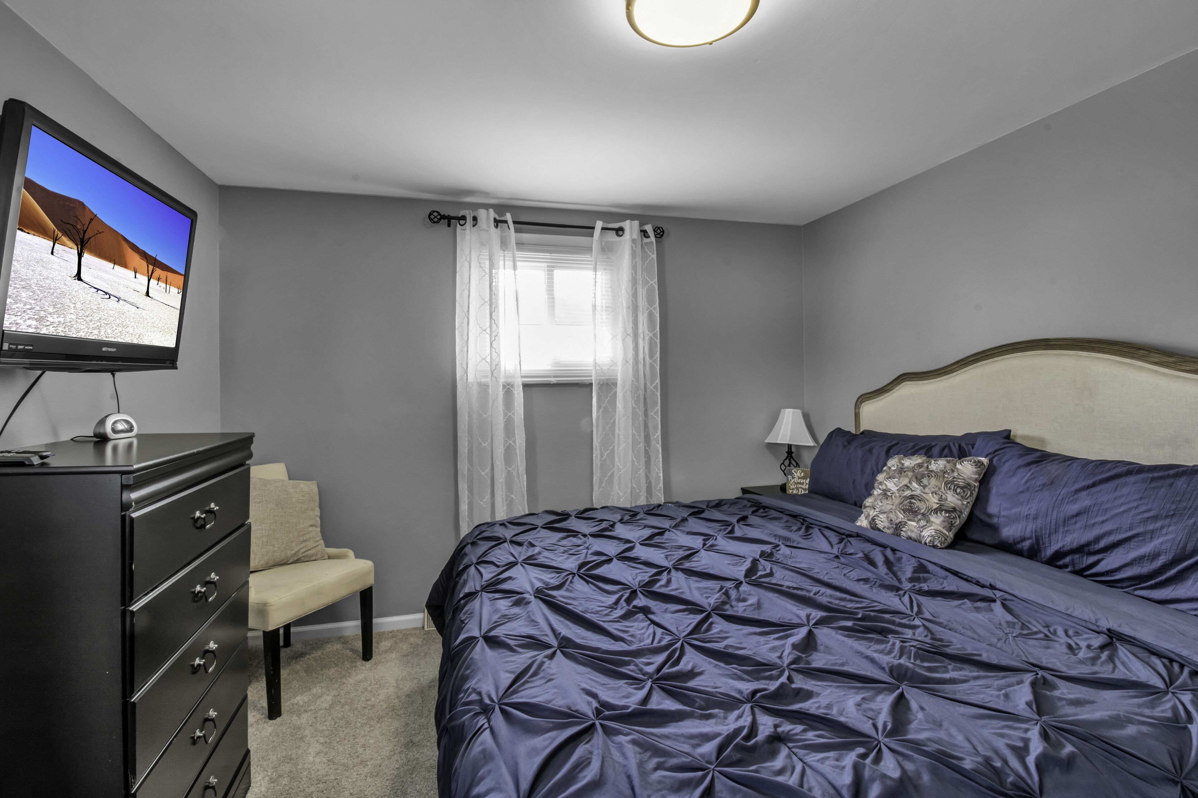 A bedroom with a bed, a dresser, a chair, a window with curtains, and a television on the wall.