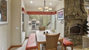 Un comedor con chimenea de piedra, una araña y vistas a una casa a través de amplios ventanales.