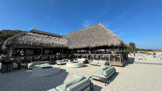Un resort frente a la playa con estructuras de techo de paja, mobiliario exterior y una playa arenosa.