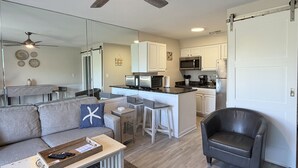 Una cocina moderna con armarios blancos, un microondas y un refrigerador. Hay un comedor con una mesa y sillas, y una sala de estar con un sofá y una mesita de centro.