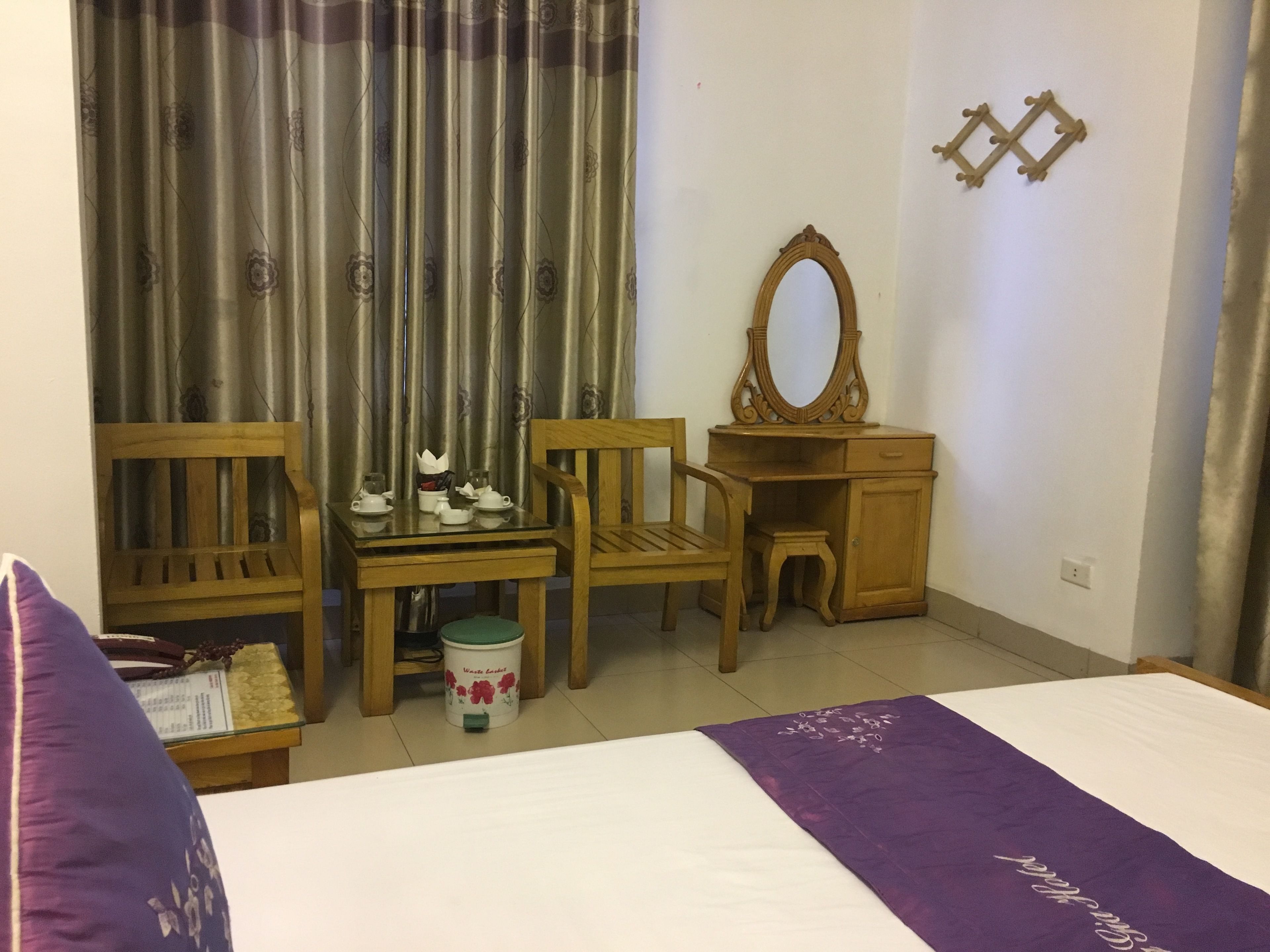 A hotel room with a bed, a wooden desk with a mirror, two chairs, a small table with a teapot, and a trash bin.