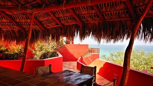 A terrace with a thatched roof, red walls, and a view of the ocean. - The Red House - Quiet, romantic weekend (Los Barriles)
