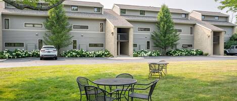 A multi-story residential building with a grassy lawn, outdoor seating area, and a car parked in front.