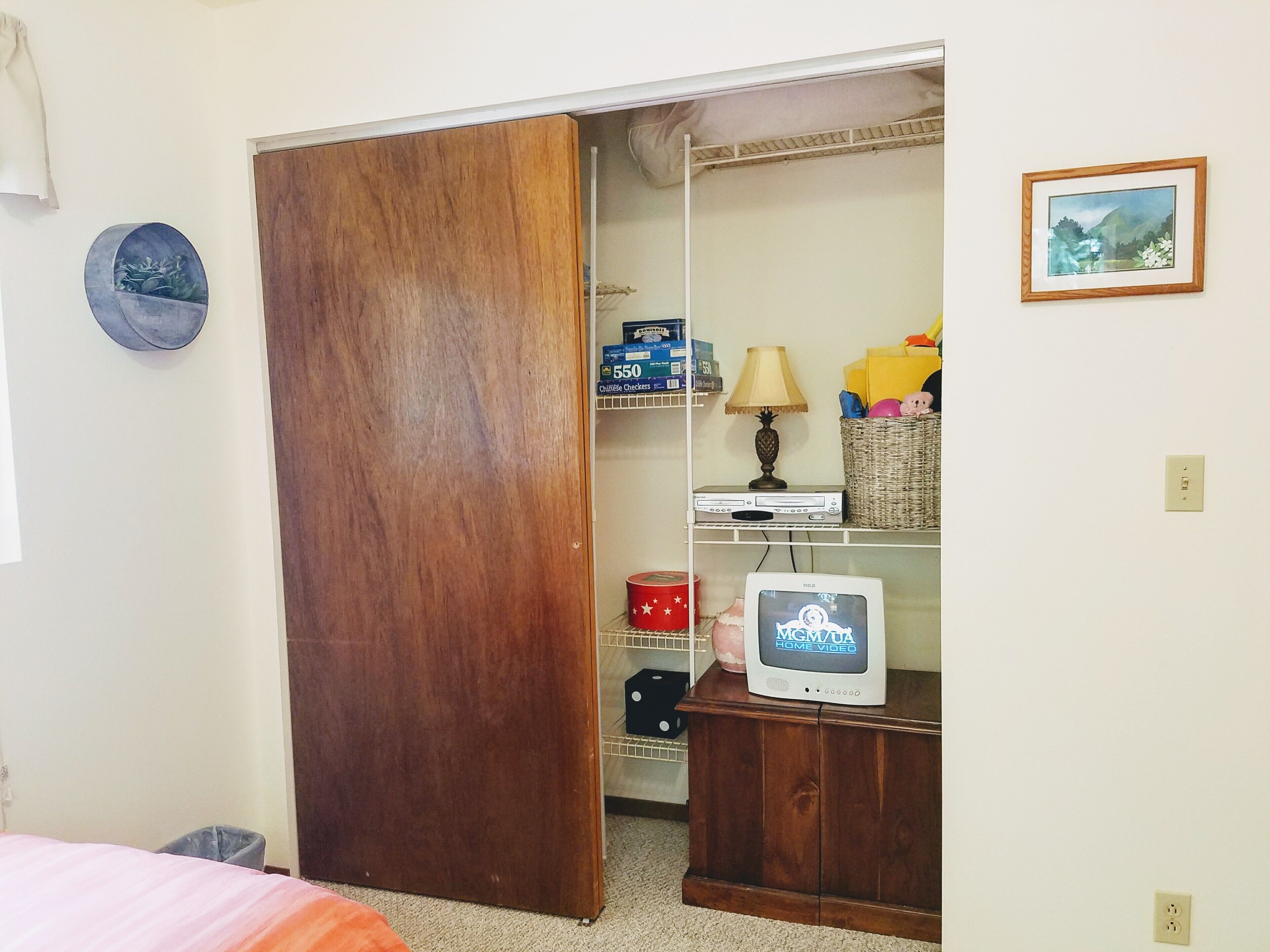 A room with a wooden wardrobe, a television on a wooden stand, a shelf with various items, and a wall-mounted picture.