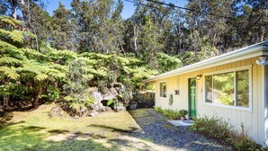Una casa con puerta verde, rodeada por un jardín con helechos y una zona rocosa.
