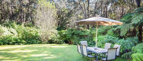 Un espace repas avec une table et des chaises sous une pergola, entouré d’une végétation luxuriante et d’arbres.