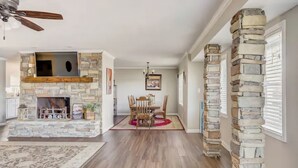 A spacious living area with a stone fireplace, wooden flooring, a dining table with chairs, and a ceiling fan.