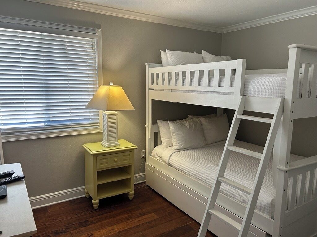 A bunk bed room with a nightstand, lamp, and a desk with a remote control.
