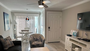 A living room with a sofa, a coffee table, a patterned armchair, a dining table with chairs, a ceiling fan, a TV, and a white cabinet. - Condo with Private Beach Access, Great Shared Amenities - Private Beach House (Siesta Key)