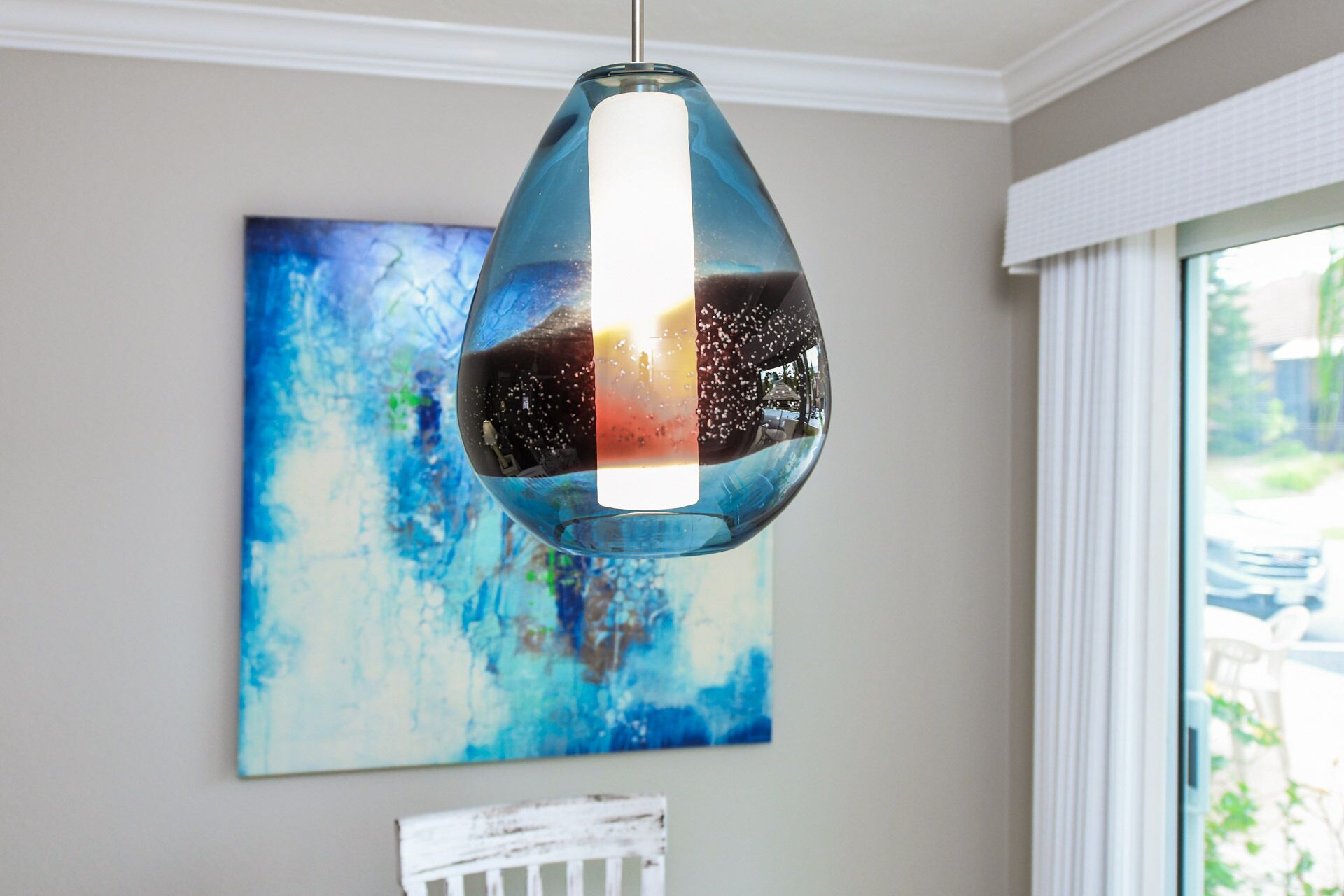 Eine moderne Pendelleuchte mit reflektierender Oberfläche, ein blaues abstraktes Gemälde an der Wand und ein weißes Geländer in der Nähe eines Fensters.