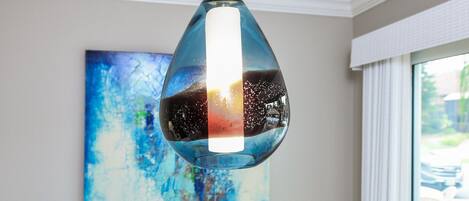 Eine moderne Pendelleuchte mit reflektierender Oberfläche, ein blaues abstraktes Gemälde an der Wand und ein weißes Geländer in der Nähe eines Fensters.