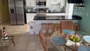A modern kitchen with white cabinets, a stainless steel refrigerator, a microwave, and a coffee maker. There is a dining area with a glass table and wicker chairs, and a small trash bin. - DUPLEX IN DUCK KEY, 2 Bedroom/2 Bath, W/Private 90' Dock,  Trailer Storage (Duck Key)