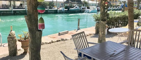 Zona junto a la piscina con mesa y sillas, rodeada de árboles y vista a barcos en el agua.