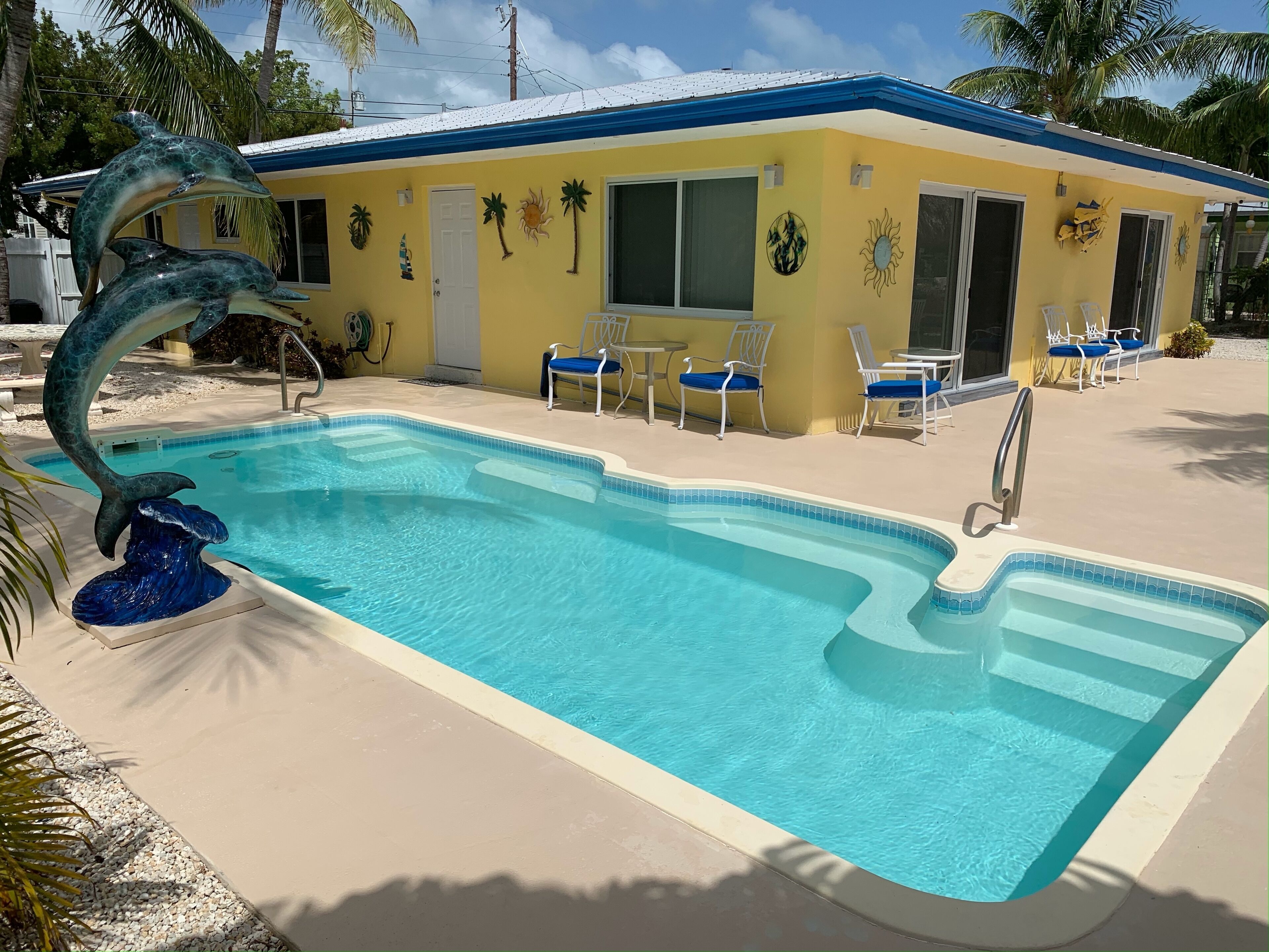 A swimming pool with a dolphin sculpture, surrounded by lounge chairs and a building with tropical wall art.
