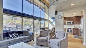 A modern living room with a sofa, armchairs, and a coffee table. A large window offers a view of a balcony and trees.