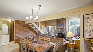 Een gezellig, landelijk interieur met een houten eettafel, een open haard en een trap naar een hoger gelegen verdieping.