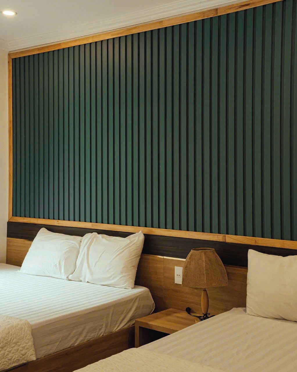 Habitación de hotel con dos camas, cabecero de madera, panel acústico verde y lámpara de noche.
