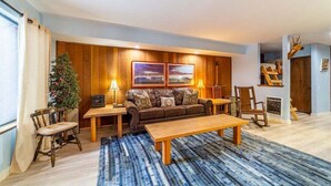 Un salon chaleureux avec un mur lambrissé en bois, un canapé en cuir marron, une table basse en bois, un tapis à rayures bleues et un petit sapin de Noël.