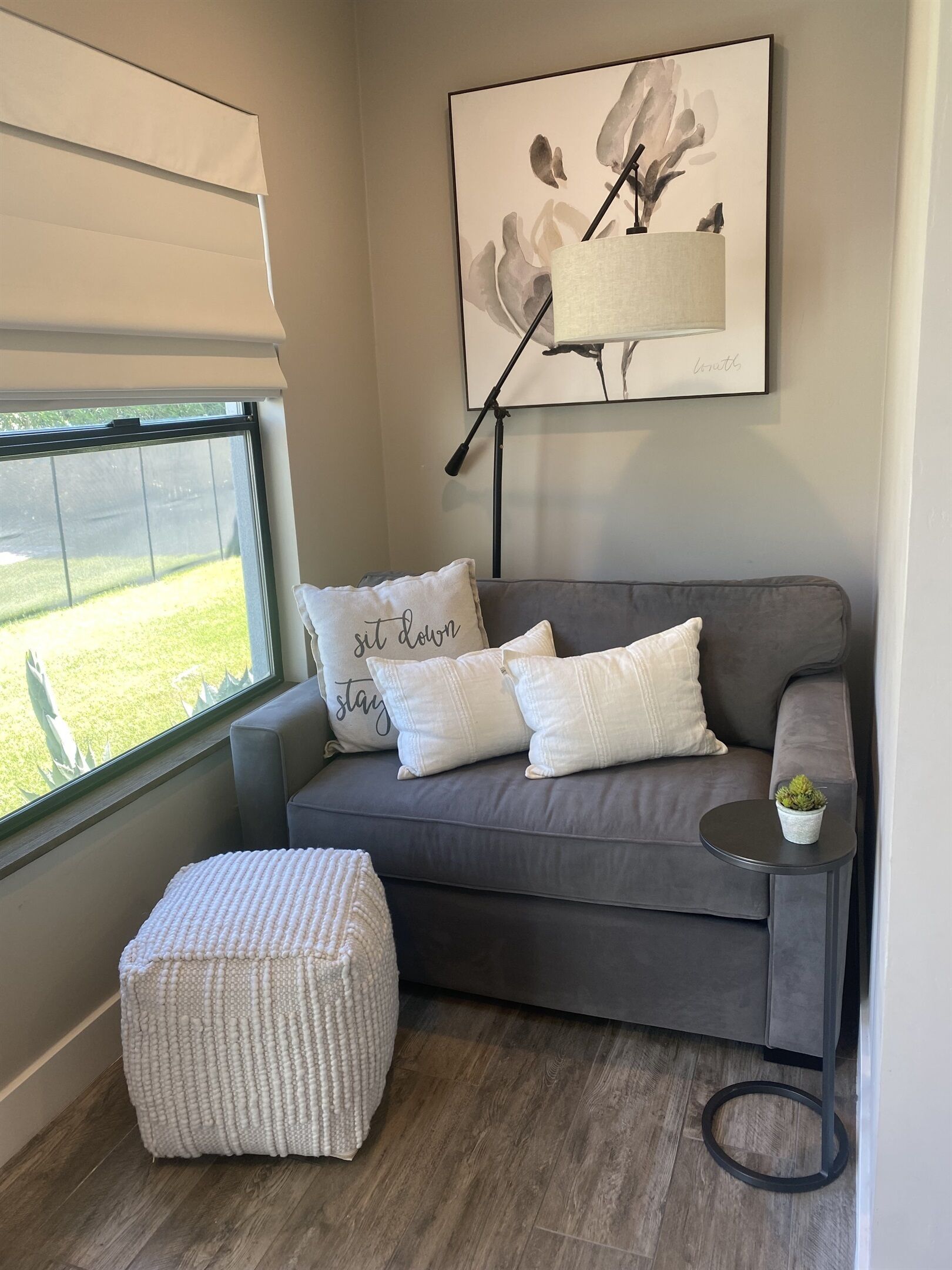 Un salon moderne avec un canapé gris, des coussins blancs, une petite table ronde ornée d’une plante et un tableau abstrait encadré accroché au mur.