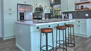A kitchen with light blue cabinets, a stainless steel refrigerator, a microwave, and a coffee maker. There are three stools at the island. - Stylish Retreat: Heated Pool w/ Jets, Putting Green & Outdoor Kitchen (Phoenix)