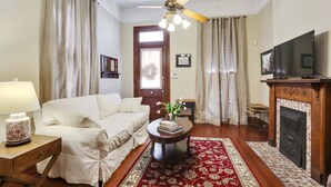 A living room with a sofa, a coffee table with books and flowers, a fireplace, and a television. - Large Shotgun Double In Perfect Uptown Location  (New Orleans)