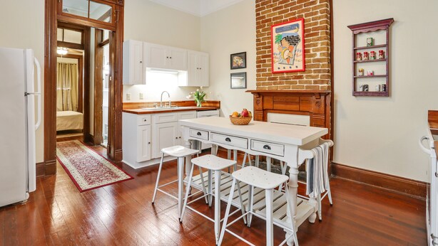 A kitchen with white cabinets, a wooden countertop, and a brick wall. There are three white stools and a white table. - Large Shotgun Double In Perfect Uptown Location  (New Orleans)