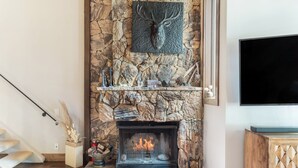 A stone fireplace with a mounted deer head, a TV above it, and a staircase to the left. - Ridgepoint 202 | Updated 3BR+Loft Ski-Way Townhome (Avon)