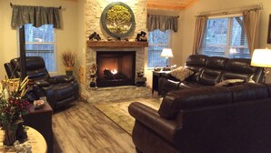 A cozy living room with a stone fireplace, leather sofas, and a large window. - Built in 2018 this is a charming, spacious chalet that sleeps 6!   (Gatlinburg)