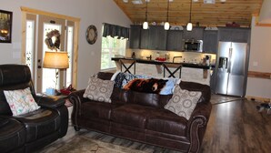 A living room with a brown leather sofa, a dining table, and a kitchen area with modern appliances. - Built in 2018 this is a charming, spacious chalet that sleeps 6!   (Gatlinburg)